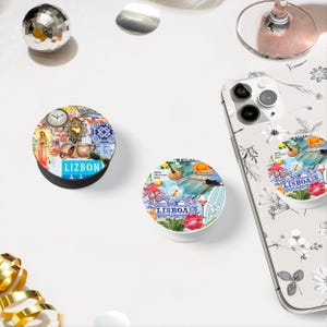 May include: Two colorful phone grips with a Lisbon theme. One grip features a collage of images including a clock, a heart, and a building. The other grip features a collage of images including a guitar, a surfboard, and a red lip. Both grips have the word "LISBOA" printed on them.
