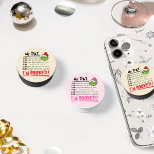 May include: Two round phone grips with a pink and beige background. Both grips feature a green Grinch character wearing a Santa hat and a list of activities for the day. The text on the grips reads "My Day" and "I'm Booked!"