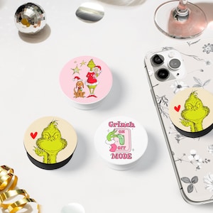 May include: Three different phone pop sockets featuring the Grinch. One is pink with the Grinch and Max the dog, one is white with the Grinch and the text "Grinch On Off Mode", and one is beige with the Grinch.