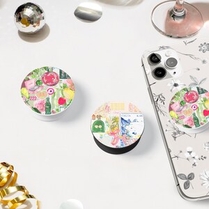 May include: Two white phone grips with colorful designs. One grip features a collage of images including a pink flamingo, a palm tree, and a vintage postcard. The other grip features a collage of images including a red and white striped beach umbrella, a bottle of soda, and a pink flower.