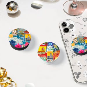 May include: Two colorful phone grips with a variety of images, including a wave, a building, a flag, and a fruit. The grips are round and have a raised edge. The grip on the left is black and the grip on the right is white.