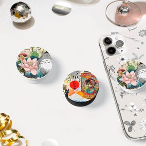Könnte beinhalten: Weiße und schwarze PopSockets mit farbenfrohen Designs. Eines zeigt eine rosa Lotusblume und eine Cartoonfigur, das andere eine Collage von Bildern mit japanischem Text. Ein Telefon mit floralem Gehäuse ist ebenfalls sichtbar.