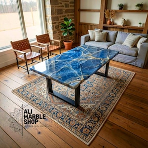 Puede incluir: Una mesa de centro rectangular con una superficie de mármol azul pulido con vetas blancas y doradas, sostenida por un marco de metal negro. La mesa está colocada sobre una alfombra estampada. El texto Ali Marble Shop es visible abajo a la izquierda.