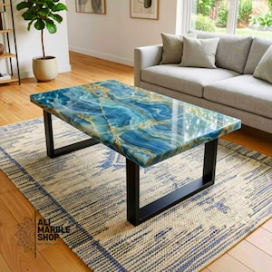 May include: A rectangular coffee table featuring a polished blue and gold marbled resin top with a glossy finish. The table is supported by two sturdy, black metal U-shaped legs. The surface displays swirling patterns of deep blue, teal, and gold veins.
