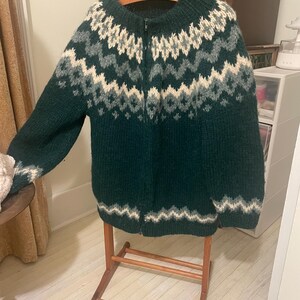 May include: A dark green zip-up sweater with a geometric pattern in white and gray. The sweater is hanging on a wooden hanger and displayed on a wooden garment rack. The sweater has long sleeves and a high neckline.