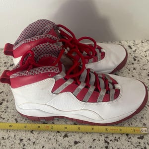 K&ouml;nnte beinhalten: Paar wei&szlig;-roter High-Top-Sneaker mit roten Schn&uuml;rsenkeln und Akzenten. Die Sneaker haben ein wei&szlig;es Lederobermaterial mit roten Zierleisten und eine rote Sohle. Zunge und Kragen weisen einen rot-wei&szlig; gemusterten Stoff auf.