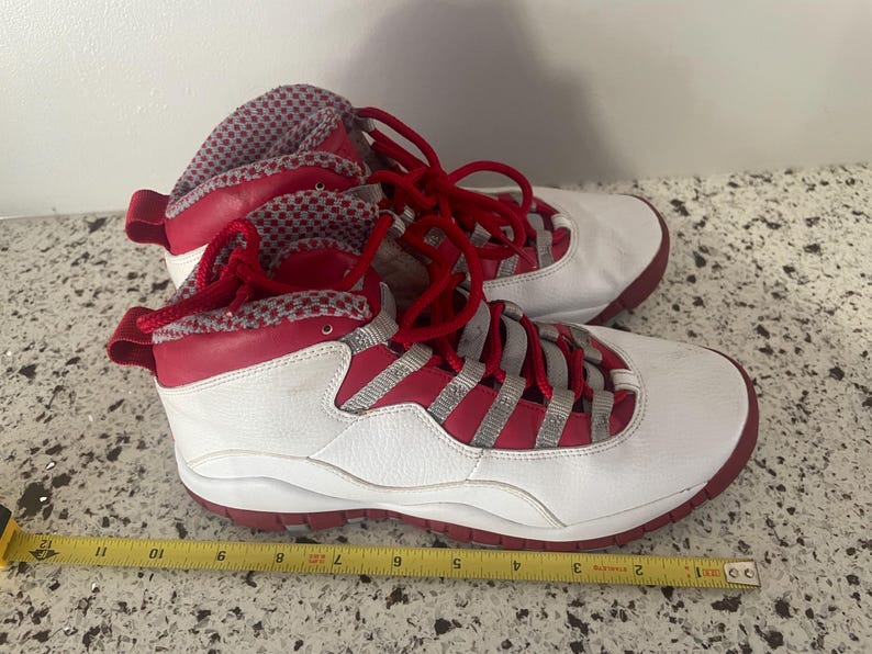 K&ouml;nnte beinhalten: Paar wei&szlig;-roter High-Top-Sneaker mit roten Schn&uuml;rsenkeln und Akzenten. Die Sneaker haben ein wei&szlig;es Lederobermaterial mit roten Zierleisten und eine rote Sohle. Zunge und Kragen weisen einen rot-wei&szlig; gemusterten Stoff auf.