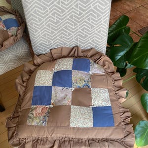 May include: A brown quilted chair cushion with a ruffled edge. The cushion is made of patchwork fabric in shades of blue, brown, and white.