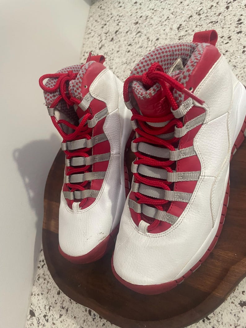 K&ouml;nnte beinhalten: Paar wei&szlig;-roter High-Top-Sneaker. Die Sneaker haben rote Schn&uuml;rsenkel, eine rote Sohle und rote Akzente an Kragen und Zunge. Die Oberteile sind wei&szlig; mit silbernen Akzenten. Die Sneaker werden auf einer Holzoberfl&auml;che pr&auml;sentiert.
