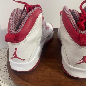 K&ouml;nnte beinhalten: Paar wei&szlig;-roter High-Top-Sneaker. Die Sneaker haben rote Akzente an Kragen, Schn&uuml;rsenkeln und Sohle, mit einem wei&szlig;en K&ouml;rper. Das Jumpman-Logo ist auf der Seite sichtbar. Der Innenkragen hat ein grau-rotes Muster.