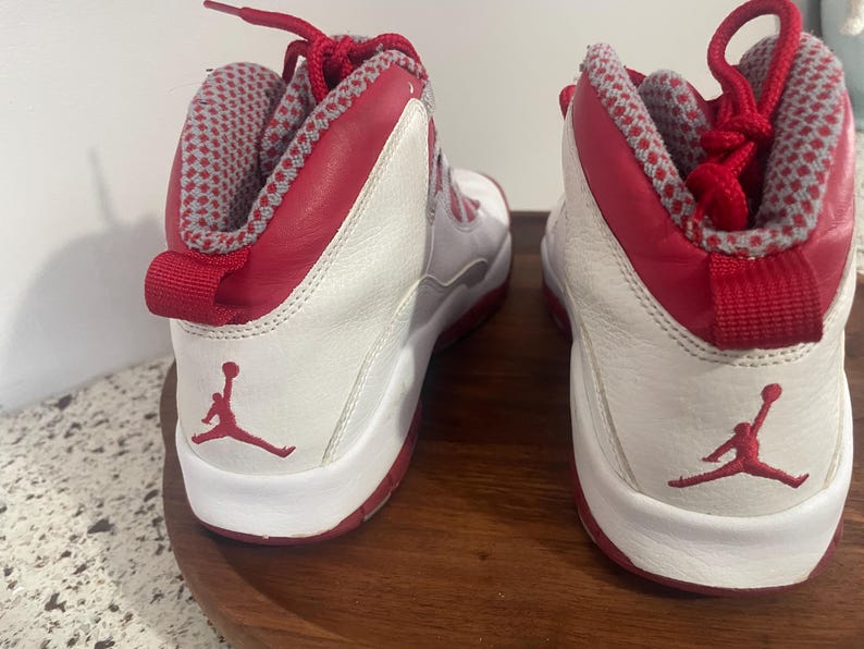 K&ouml;nnte beinhalten: Paar wei&szlig;-roter High-Top-Sneaker. Die Sneaker haben rote Akzente an Kragen, Schn&uuml;rsenkeln und Sohle, mit einem wei&szlig;en K&ouml;rper. Das Jumpman-Logo ist auf der Seite sichtbar. Der Innenkragen hat ein grau-rotes Muster.