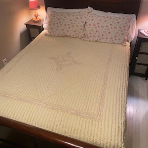 May include: A cream-coloured, tufted bedspread with a diamond pattern and a ruffled border. The bedspread is on a wooden bed frame with a headboard and footboard. Two pink floral patterned pillows are on the bed.