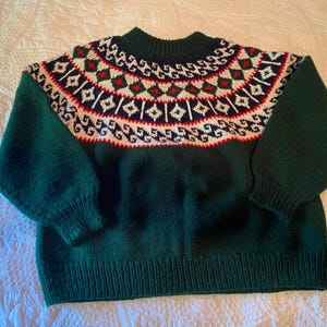 Könnte beinhalten: Ein grüner Strickpullover mit einem weißen, roten und blauen geometrischen Muster um den Hals.