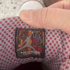 K&ouml;nnte beinhalten: Nahaufnahme des Innenraums eines rot-wei&szlig;en Air Jordan Sneakers. Die Innensohle weist ein rot-graues Karomuster auf. Ein schwarzes und goldenes Etikett tr&auml;gt die Aufschrift "AIR JORDAN COLLECTION" mit dem ikonischen Jumpman-Logo.