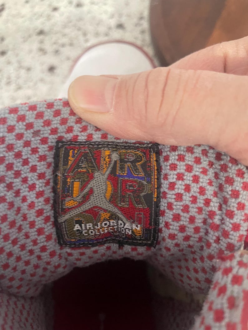 K&ouml;nnte beinhalten: Nahaufnahme des Innenraums eines rot-wei&szlig;en Air Jordan Sneakers. Die Innensohle weist ein rot-graues Karomuster auf. Ein schwarzes und goldenes Etikett tr&auml;gt die Aufschrift "AIR JORDAN COLLECTION" mit dem ikonischen Jumpman-Logo.