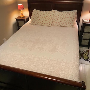 May include: A white bed with a floral patterned bedspread and two floral patterned pillows. The bed is made of wood and has a dark brown headboard. There are two bedside tables with lamps on either side of the bed.