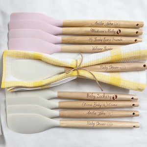 May include: A collection of personalized spatulas with wooden handles and silicone heads in pink and white. The spatulas are engraved with names and phrases like "Baby Shower" and "Welcome Sindy". They are bundled with checkered kitchen towels.