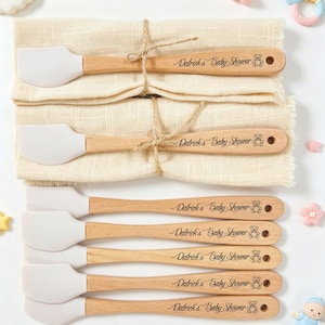 Puede incluir: Un conjunto de espátulas de silicona blanca con mangos de madera, cada una grabada con "Patrick's Baby Shower" y un icono de oso de peluche. Las espátulas están atadas con hilo y presentadas sobre una tela beige, rodeadas de decoraciones para bebés.