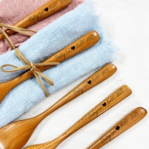 Könnte beinhalten: Hellbraune Holzlöffel mit der Gravur "Baby W ❤ EST 2025". Sie liegen auf rosa und blauen Stoffen und sind mit einem Lederband gebunden. Die Löffel sind für ein Baby.