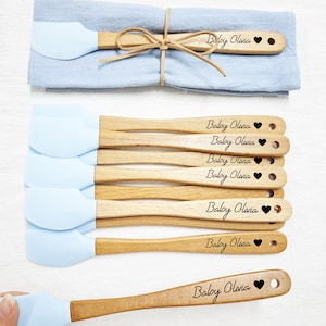 May include: A set of light blue silicone spatulas with wooden handles, each engraved with "Baby Olivia" and a heart. The spatulas are arranged on a white surface, with a blue cloth and a spatula tied with twine in the background.