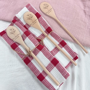 Puede incluir: Tres cucharas de madera con diseños florales grabados y texto, atadas con hilo, descansan sobre un paño de cocina a cuadros rojos y blancos. Una cuarta cuchara se coloca por separado. El texto en las cucharas dice "Emma's Bridal Shower 06.21.2026".