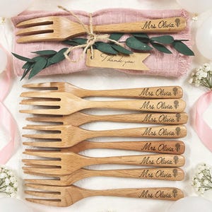 May include: Wooden forks with engraved text "Mrs. Olivia" and a floral design. The forks are arranged on a pink cloth, with a "Thank you" tag and greenery. A set of eight forks are displayed.