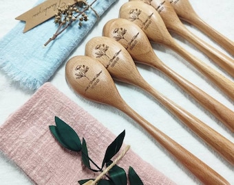 May include: Wooden spoons with engraved floral designs and text. The spoons are arranged on a white surface with blue and pink linen napkins, dried flowers, and a tag that says "Thank you".
