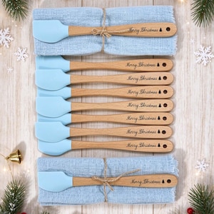 Puede incluir: Un juego de ocho espátulas de cocina con cabezales de silicona azul claro y mangos de madera. Cada mango está grabado con el texto Merry Christmas y un pequeño icono de árbol. Las espátulas están dispuestas sobre dos servilletas de tela azul claro.
