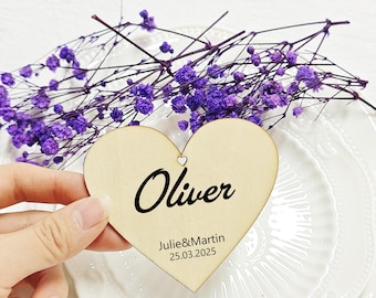 May include: A wooden heart-shaped place card with the name "Oliver" in black script. Below, the names "Julie&Martin" and the date "25.03.2025" are printed. The card is held over a white plate and decorated with purple flowers.