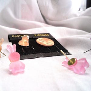 May include: A collection of bunny-shaped fashion earrings in various colors, including pink, peach, and purple. The earrings are displayed on a black earring display card with the words "FASHION EARRINGS" in gold lettering. A penny is also on the display.