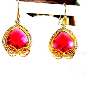 May include: A pair of gold-tone drop earrings featuring faceted heart-shaped red stones. Each earring is framed by a textured gold-tone border with a small decorative bow detail at the base and hangs from a simple gold-tone hook wire finding.