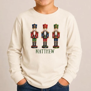 May include: A cream-colored long-sleeve shirt featuring a graphic of three nutcracker figures in red, blue, and green uniforms. Below the nutcrackers, the name MATTHEW is printed in green capital letters. The shirt has a crew neckline.