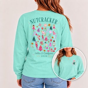 May include: A light teal long-sleeve shirt featuring a back graphic with the text "NUTCRACKER Soldier & Candycane 2026" surrounded by pink and red holiday illustrations of nutcrackers, trees, stars, and candy. The front chest has a small circular logo.