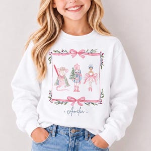 May include: A white long-sleeve sweatshirt featuring a watercolor-style graphic of a mouse, a nutcracker, and a ballerina inside a pink ribbon frame. The name Amelia is printed in cursive script below the illustration on the front.