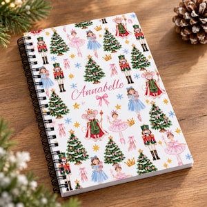 May include: A spiral-bound notebook with a white cover featuring a repeating pattern of Christmas trees, nutcrackers, ballerinas, mice in royal attire, crowns, bows, and snowflakes. The name Annabelle is printed in pink script in the center.