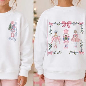 May include: A white long-sleeve sweatshirt featuring a front graphic of a nutcracker with the text "Hailey" and "Nutcracker 2026". The back displays a rectangular frame of greenery and pink bows containing illustrations of a ballerina, a nutcracker, and a girl.