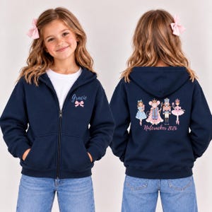 May include: A navy blue zip-up hoodie featuring a white shirt underneath. The front displays the name Gracie with a pink bow. The back features a graphic of a girl, a mouse, a nutcracker, and a ballerina with the text Nutcracker 2026.