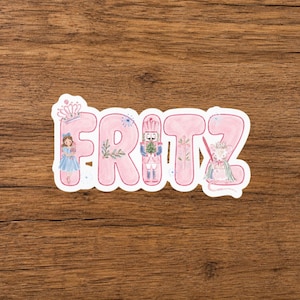 May include: A die-cut sticker featuring the name FRITZ in large, light pink, hand-painted style letters. The letter F shows a girl in a blue dress, the R contains greenery, the I features a nutcracker, the T shows a sword, and the Z a mouse.