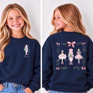 May include: A navy blue sweatshirt featuring a small nutcracker graphic with the text Nutcracker 2024 and the name Charlie on the front left chest. The back displays a larger illustration of a ballerina, a nutcracker, and a girl.
