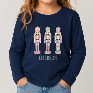 May include: A navy blue long-sleeve shirt featuring a graphic of three pastel-colored nutcrackers arranged in a row. Below the nutcrackers, the name EMERSON is printed in a light green, serif font. The shirt has a crew neckline and long sleeves.