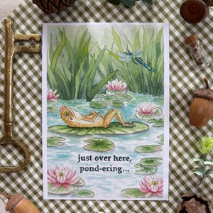 Könnte beinhalten: Grußkarte mit einer Aquarellillustration eines Frosches, der sich auf einer Seerose in einem Teich entspannt. Die Karte trägt den Text "just over here, pond-ering...". Die Szene umfasst Seerosen und eine Libelle.