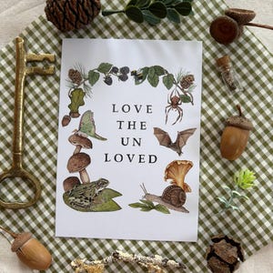 May include: A white print with the text "LOVE THE UNLOVED" surrounded by illustrations of a frog, snail, mushrooms, a bat, and a spider. The print is on a green and white checkered surface, with decorative acorns, a key, and pinecones.