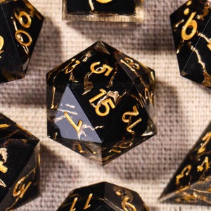Puede incluir: Un juego de siete dados poliédricos con base negra, vetas doradas y números dorados. El conjunto incluye un d20, d12, d10, d8, d6, d4 y un dado de porcentaje, todos ellos con números claros y legibles en una fuente estándar.