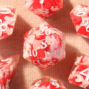 May include: A set of seven polyhedral dice, each with red and pink heart-shaped inclusions and white numbers. These dice are designed for role-playing games. The dice are displayed on a light pink surface.