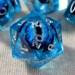 Puede incluir: Un conjunto de dados poliédricos de resina azul translúcida. Cada dado presenta un ojo azul realista y detallado con una pupila vertical negra incrustada en el centro. Los números de los dados están grabados y rellenos de pintura plateada.
