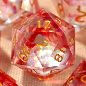 Puede incluir: Un conjunto de dados poliédricos con un diseño transparente, rojo y dorado. Los dados son transparentes con remolinos rojos y numeración dorada. El conjunto incluye varias formas, como un dado de 20 caras, y probablemente se utiliza para juegos de mesa.