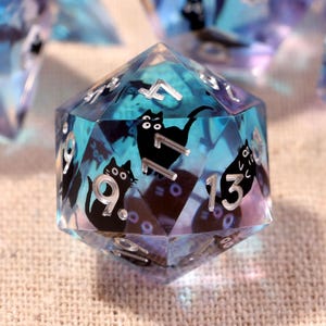May include: A set of polyhedral dice made from translucent blue and purple resin. Each die features embedded black cat silhouettes and silver-colored numbers. The dice have sharp edges and a glossy finish, with various shapes including twenty-sided, twelve-sided, and eight-sided forms visible in the collection.