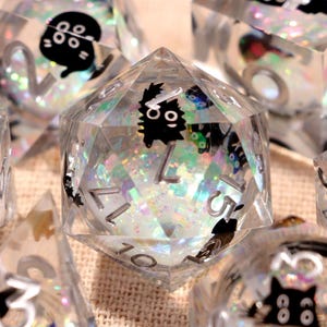 Puede incluir: Juego de siete dados de resina transparente con ilustraciones de gatos negros y purpurina iridiscente. Los dados incluyen un D4, D6, D8, D10, D12, D20 y un dado de porcentaje. Cada dado tiene números blancos.