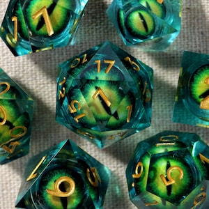 May include: A set of seven polyhedral dice with a teal and green color scheme. Each die features a detailed eye design in the center, with gold-colored numbers. The dice are arranged on a neutral-colored surface.