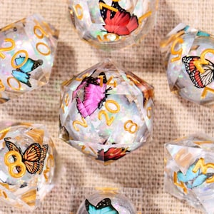 May include: A set of seven polyhedral dice made of clear resin with embedded colorful butterfly illustrations and iridescent glitter. Each die features gold-colored numbers. The set includes various shapes such as a d20, d12, d10, d8, d6, and d4.